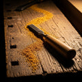 Golden sawdust shimmering in the light on a dark wood workbench next to a chisel.