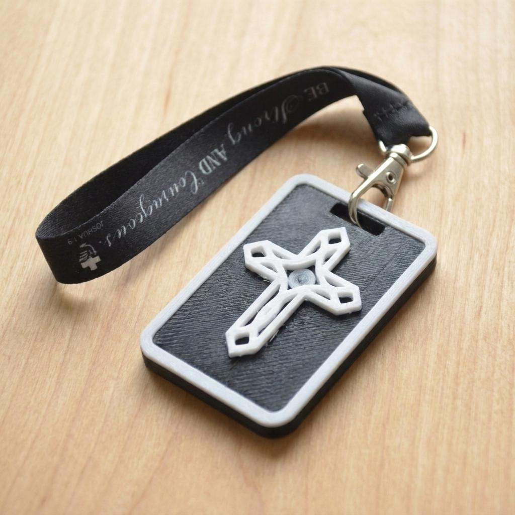 A 3D-printed Eben Tag in Black featuring a raised ornate cross design. This NFC-enabled spiritual touchpoint includes a white border and is attached to a fabric lanyard printed with scripture, designed to trigger the Daily Eb devotional.