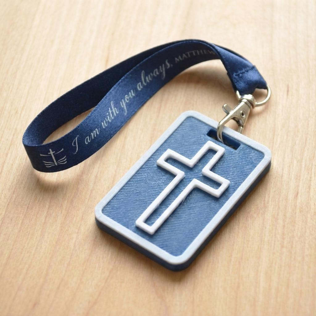 A 3D-printed Eben Tag in Carbon Blue featuring a raised cross design. This NFC-enabled spiritual touchpoint includes a white border and is attached to a fabric lanyard printed with scripture, designed to trigger the Daily Eb devotional.