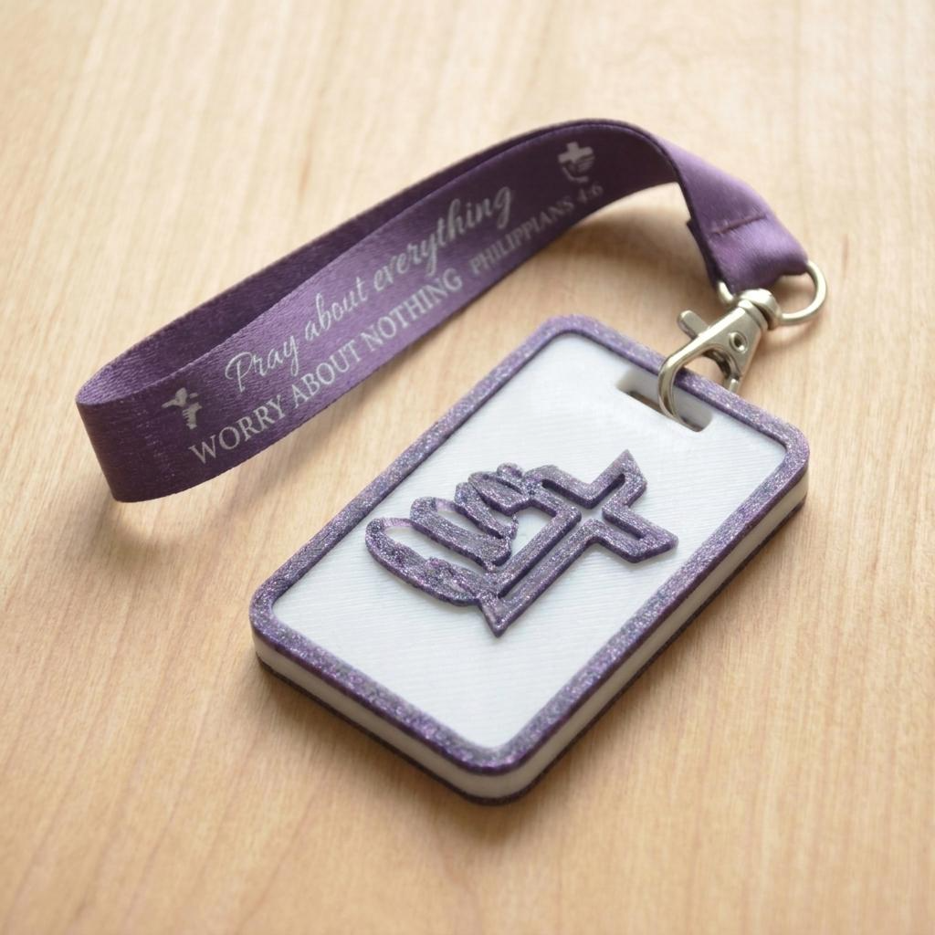 A 3D-printed Eben Tag in white and purple featuring a raised galed design. This NFC-enabled spiritual touchpoint includes a white border and is attached to a fabric lanyard printed with scripture, designed to trigger the Daily Eb devotional.
